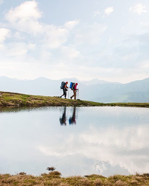 Two hikers passing by a calm mountain lake reflecting the sky